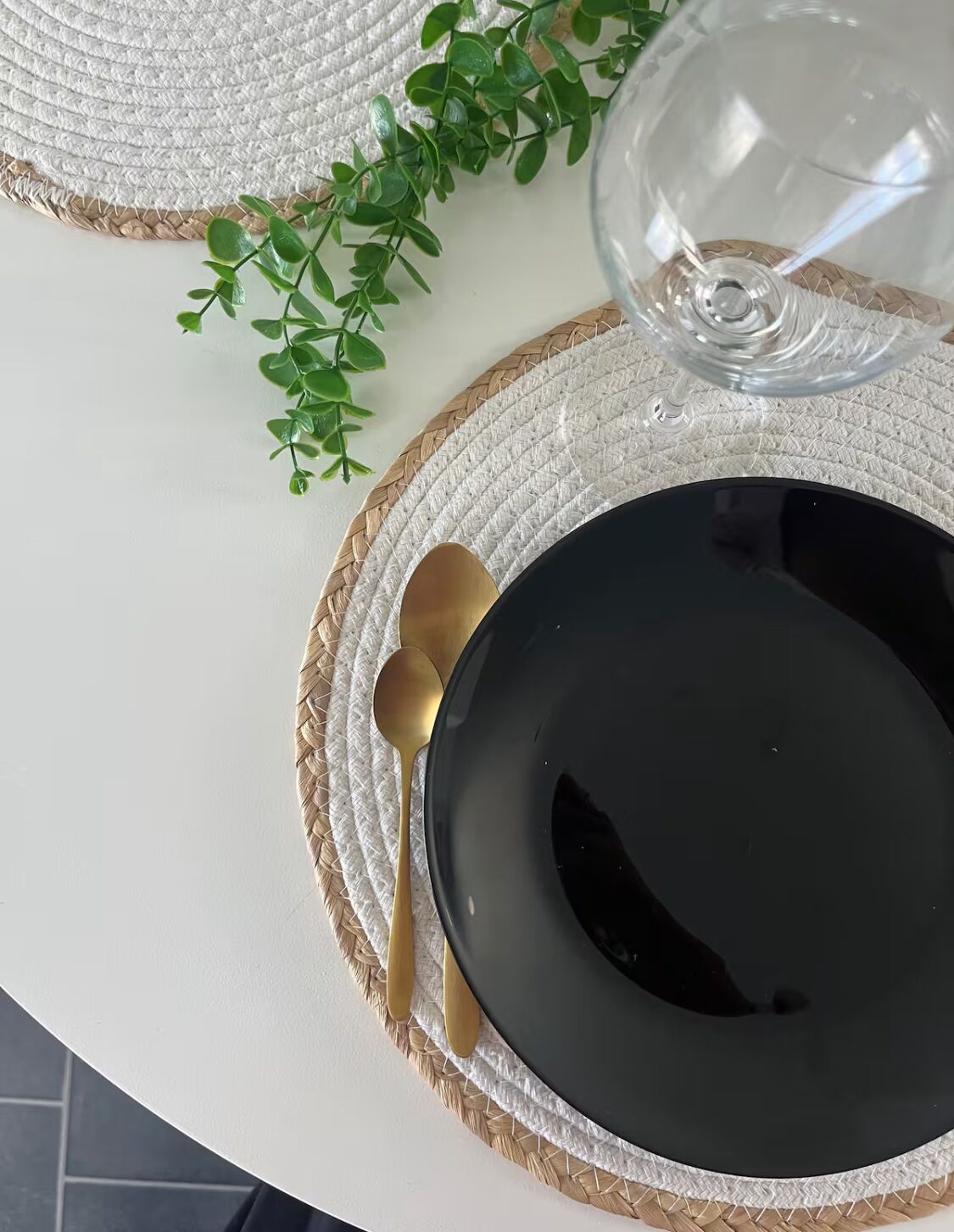 A table setting with a black plate, a gold spoon, and a glass on a woven placemat.