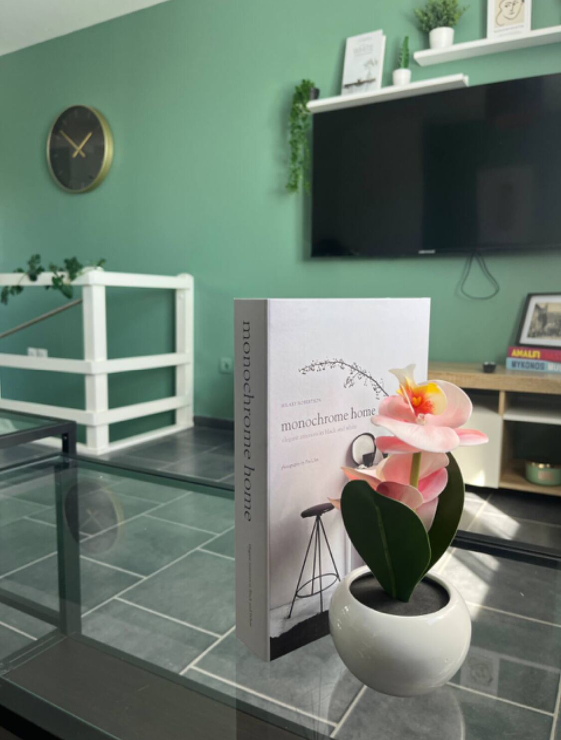Un libro con el título 'monochrome home' sobre una mesa de vidrio con una orquídea en maceta. En el fondo, una estantería blanca con objetos decorativos y un reloj.