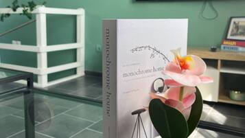Un libro con el título 'monochrome home' sobre una mesa de vidrio con una orquídea en maceta. En el fondo, una estantería blanca con objetos decorativos y un reloj.