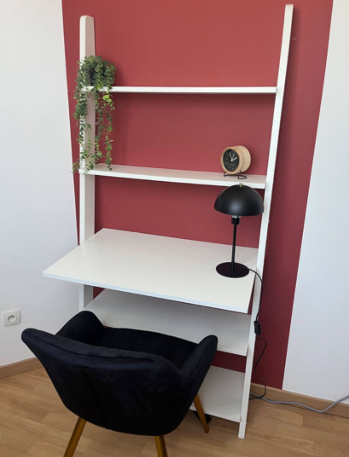 A white desk with a black chair, a clock, and a plant on a red wall.