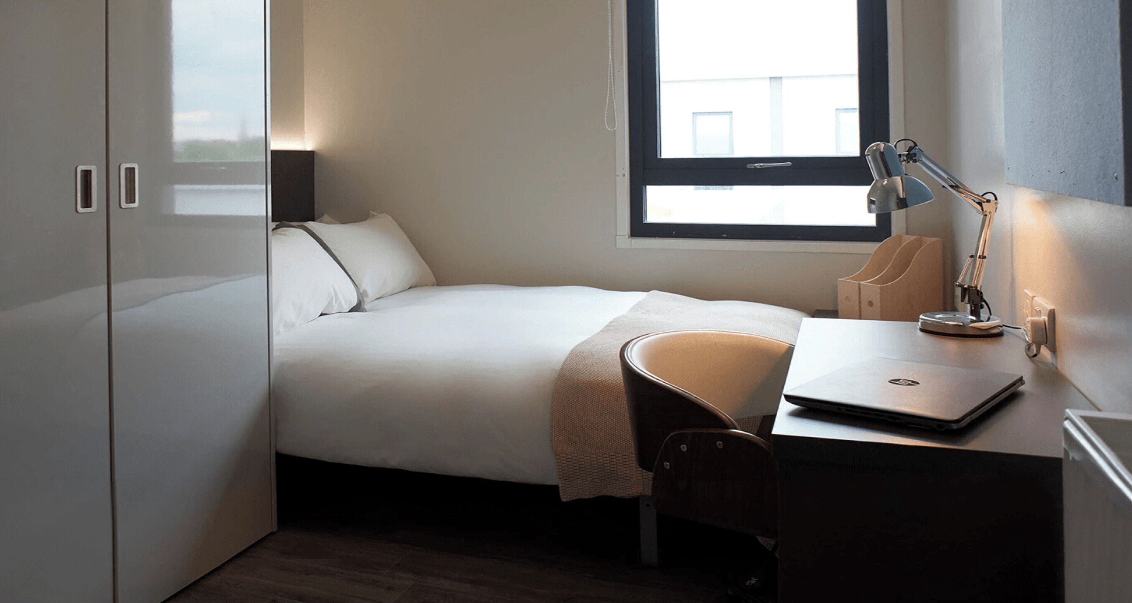 A bedroom with a bed, a desk with a laptop, a chair, and a window.