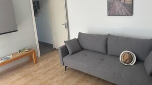 Un salon avec un canapé gris, un banc en bois, un chapeau, un tableau représentant un coucher de soleil et une porte donnant sur une autre pièce.