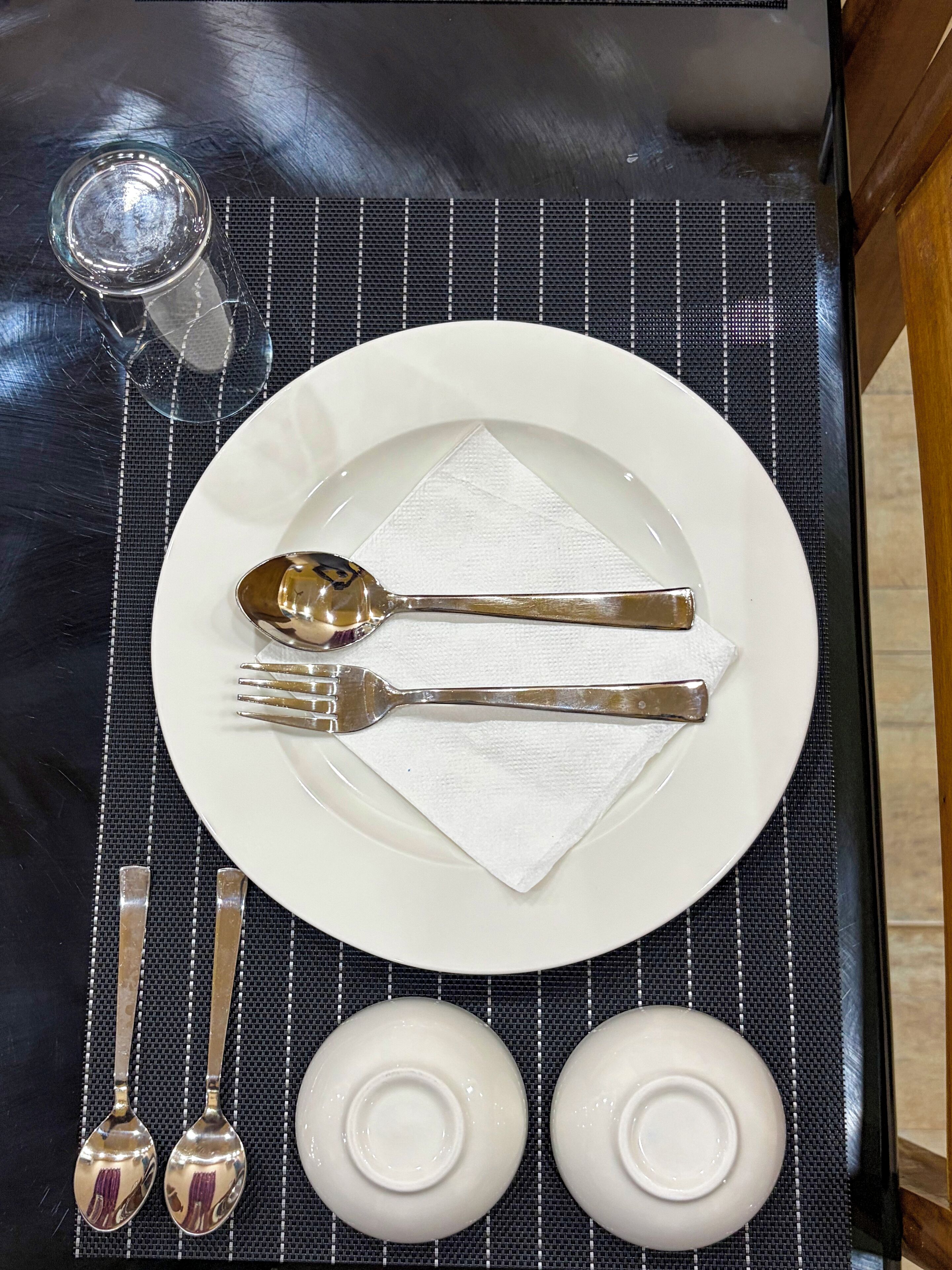 Una mesa de comedor bien puesta con un plato blanco, una cuchara, un tenedor y dos boles blancos pequeños.