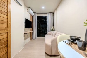 Ein modernes Wohnzimmer mit einer Couch, einem Esstisch mit Schüssel und Besteck, einem an der Wand befestigten Fernseher und einer Glastür, die auf einen Balkon führt.