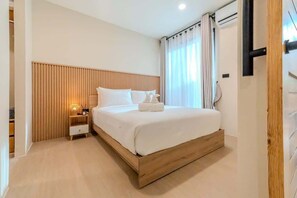 Ein modernes Hotelzimmer mit einem Holzkopfteil, einem Bett mit weißen Leinen, einem Nachttisch mit Lampe und einem Fenster mit Vorhängen.