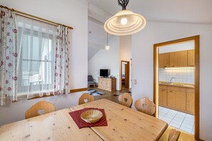 A dining area with a wooden table, chairs, a television in the background, and a kitchen visible through an open doorway. - Apartment 'Fewo Brünnstein - Room 55' with Mountain View, Garden and Wi-Fi (Oberaudorf)