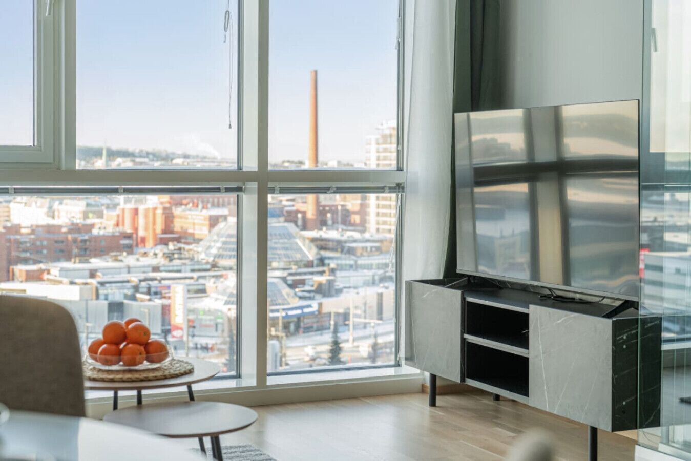 A modern living room with a large window, a flat-screen TV, and a small table with oranges.