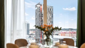 A dining table set for six with a vase of flowers, large windows offering a city view. - Topaasi - Nokia Arena by Come in (Tampere)