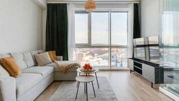 A modern living room with a sofa, a small table with fruit, a TV, and a large window with a city view.
