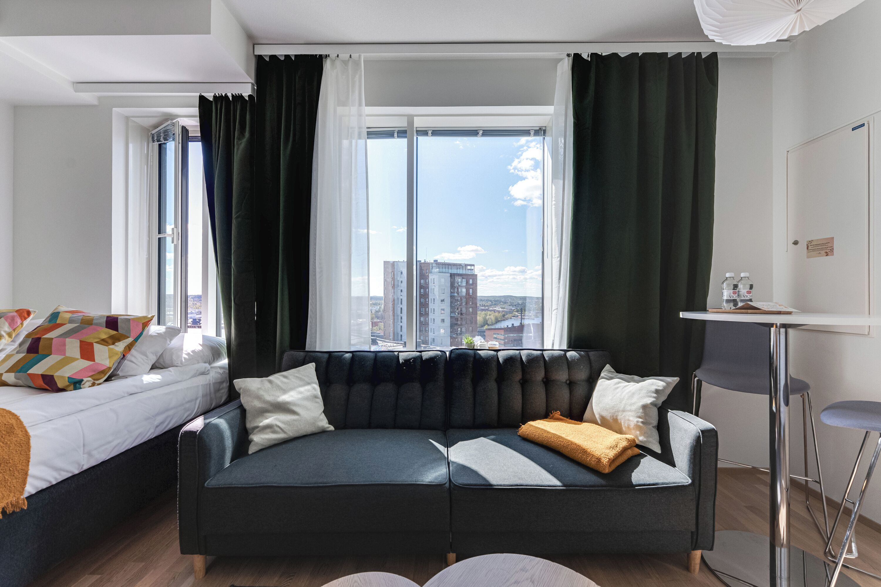 A modern hotel room with a large window, a sofa, a bed, and a small table.