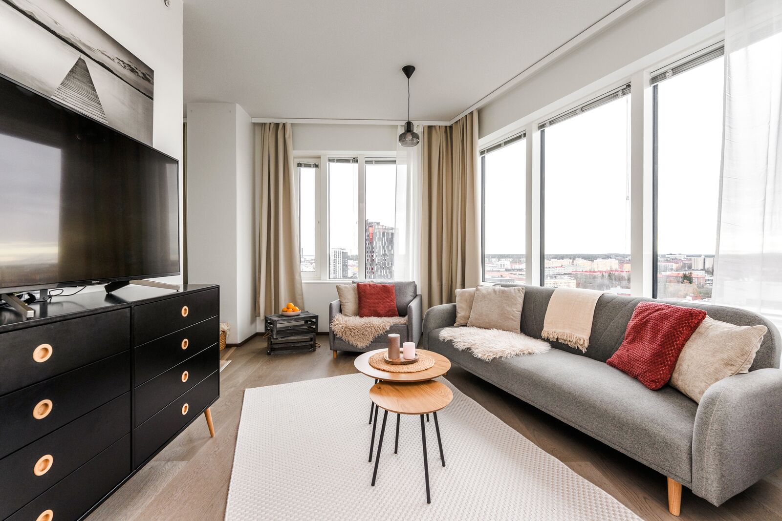 A modern living room with a grey sofa, black entertainment unit, and large windows offering a city view.