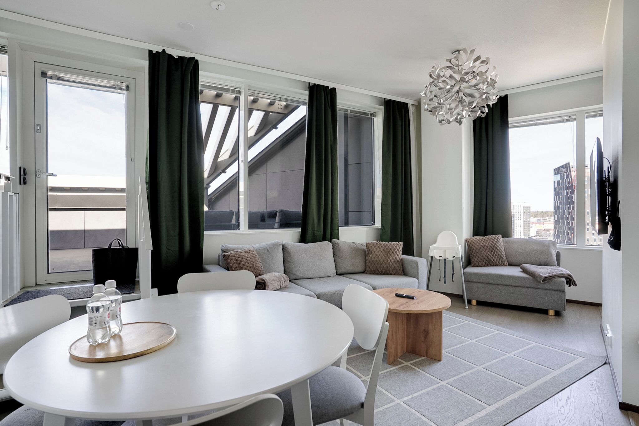 A modern living room with a white round table, grey sofa, and a city view through large windows.