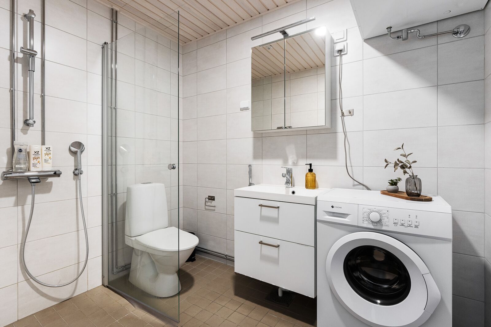Moderni kylpyhuone, jossa on lasiseinällä varustettu suihku, wc, peilillä varustettu pesuallas ja sisäänrakennettu pesukone.