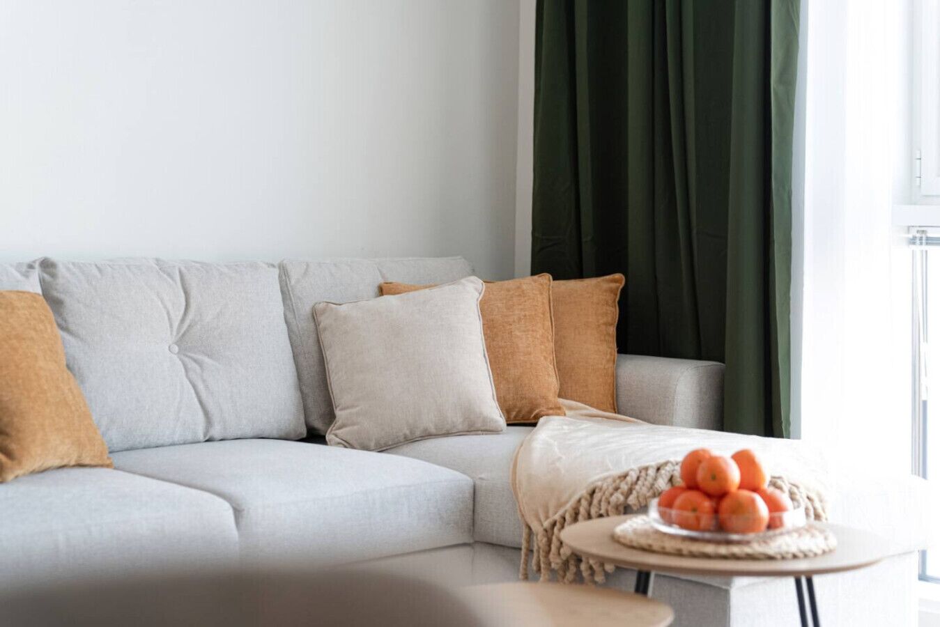 Un salon moderne avec un canapé gris, une petite table ronde ornée d’oranges et un rideau vert.
