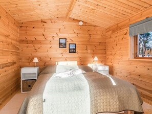 Ein Schlafzimmer einer Holzhütte mit einem Bett, zwei Nachttischen, einem Fenster und zwei gerahmten Bildern an der Wand.