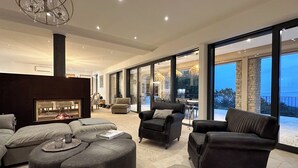 A modern living room with a fireplace, leather sofas, ottomans, and large windows offering a view of the outdoors. - Mas La Rose Sauvage – Elegant Stone Villa with Panoramic Views near Gordes (Murs)