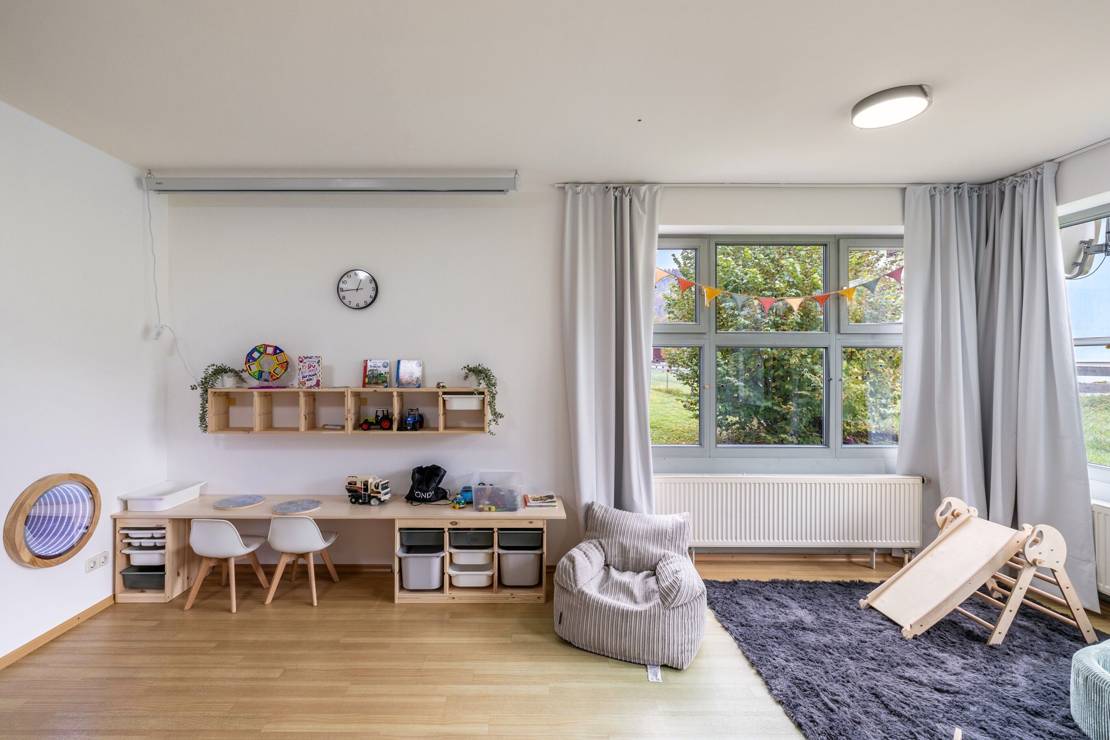 Un cuarto de juegos infantil con un escritorio de madera, sillas, un sillón de peluche, un tobogán y una ventana con cortinas.