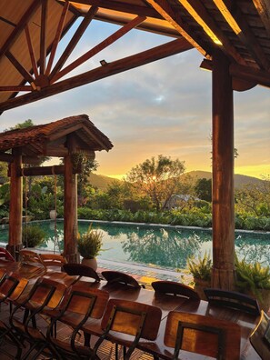 A covered outdoor seating area with a pool and chairs, surrounded by lush greenery and a view of the sunset over a hilly landscape. - LeeHa Cottage Soc Sơn (Hanoi)
