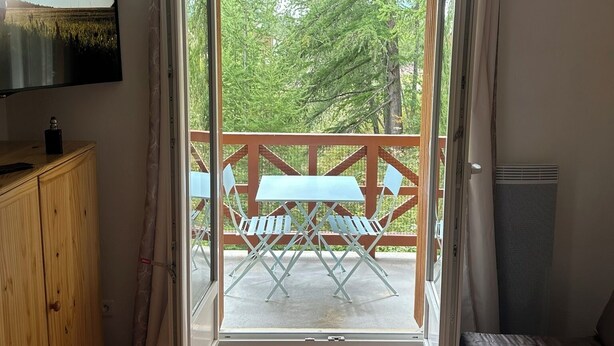 A room with a wooden floor, a bed, a TV on the wall, and an open door leading to an outdoor area with a table and chairs. - South-facing studio with direct runway access sleeps 4 Vallée Blanche residence (Allos)