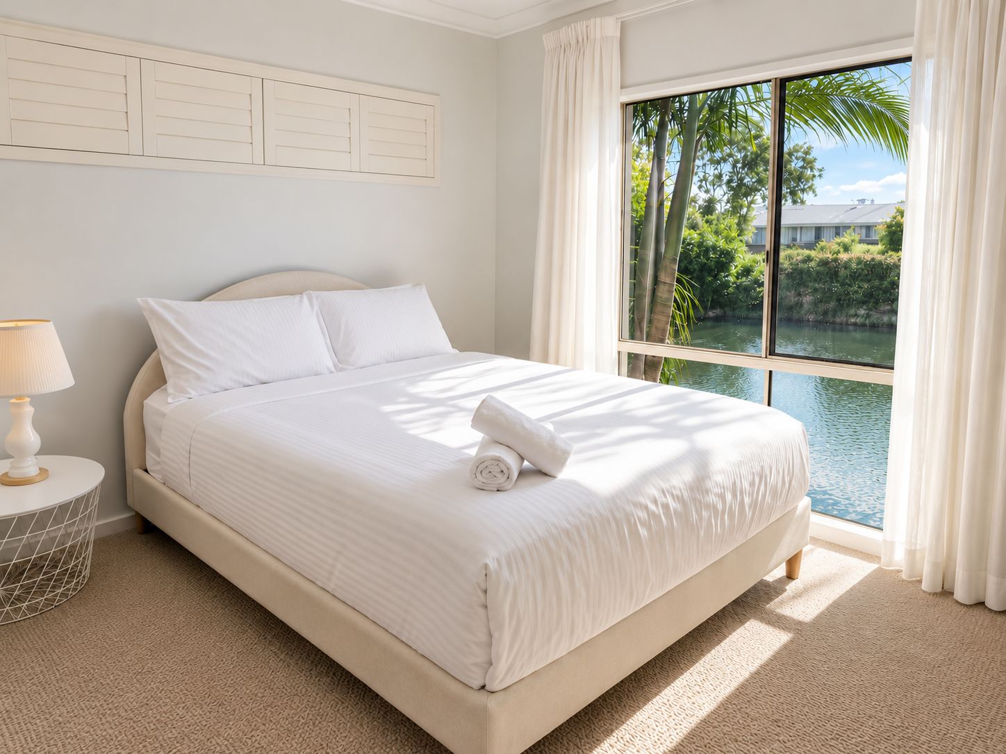 A hotel room with a large bed, a nightstand, a lamp, and a view of a body of water through a window.