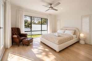 A bedroom with a large bed, a chair, a ceiling fan, and a window with a view of trees.