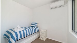 Et lille, pænt værelse med en enkelt seng, et natbord, en skuffe-kommode og en aircondition-enhed monteret på væggen.