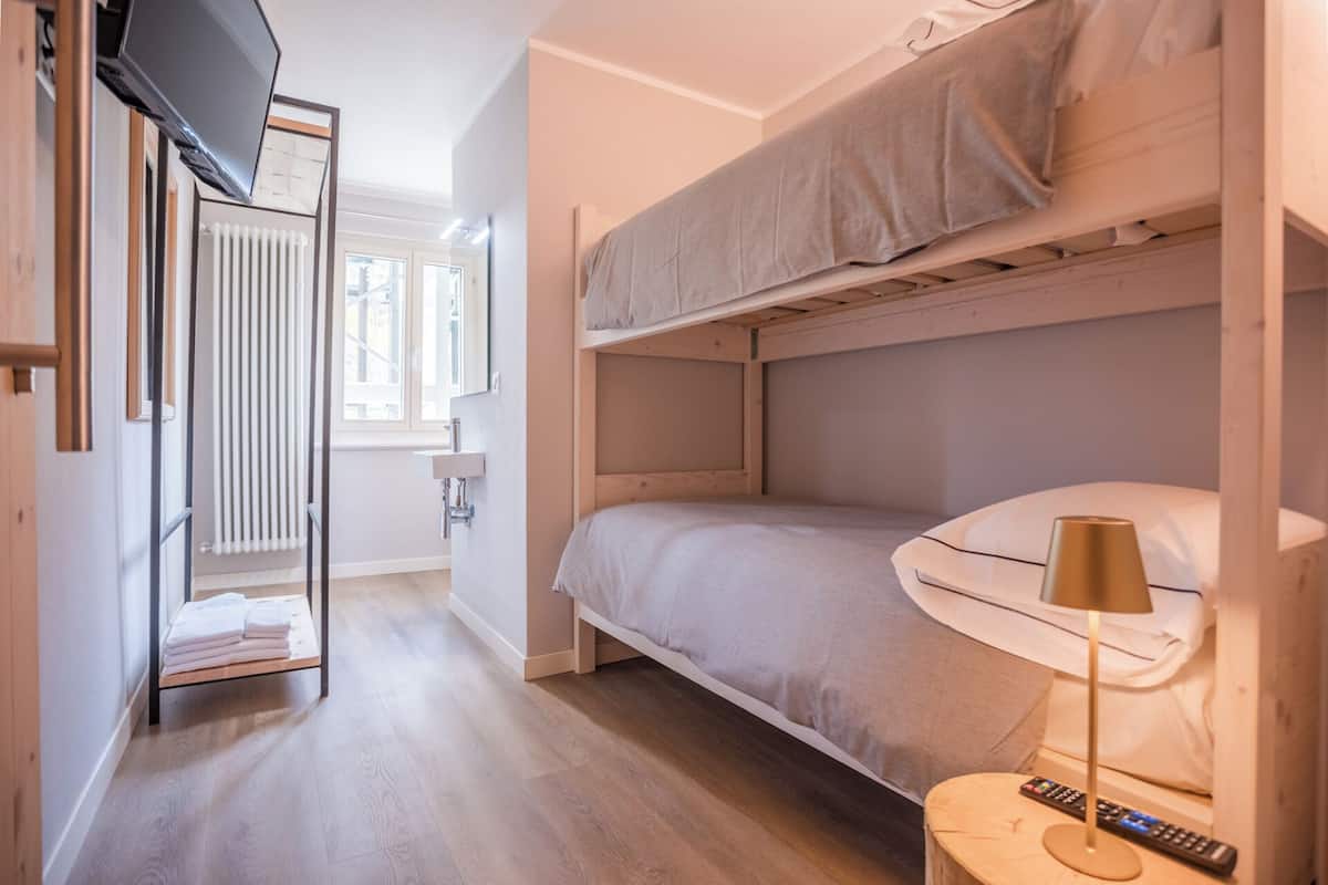 A compact room with a bunk bed, a bedside table with a lamp, a TV mounted on the wall, and a window with blinds.