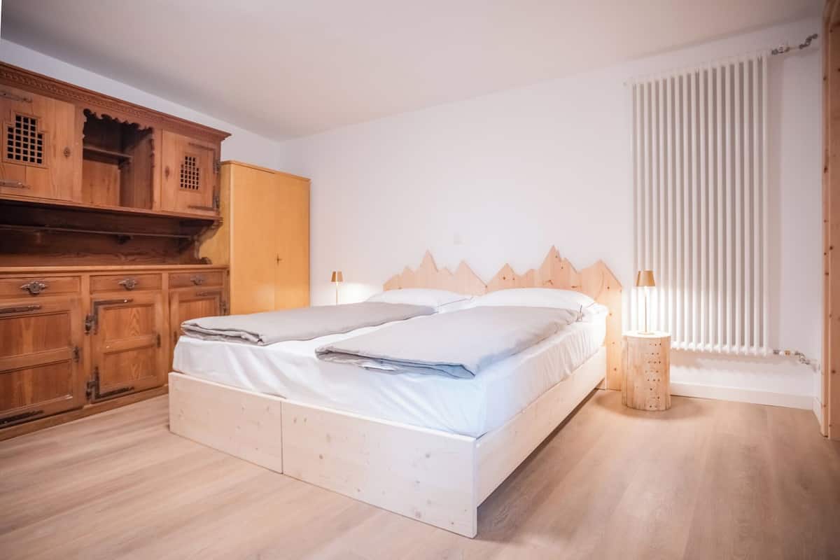 A bedroom with a wooden wardrobe, a bed with white bedding, and a window with vertical blinds.