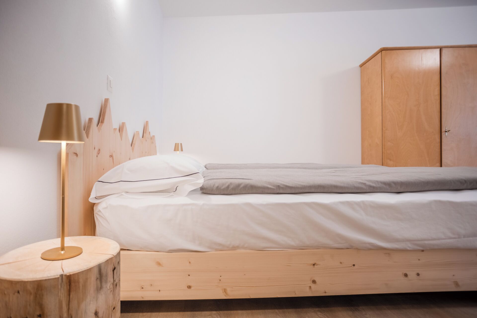 Un dormitorio con cabecera de madera, cama con ropa de cama blanca y gris, mesita de noche redonda de madera y un armario de madera.