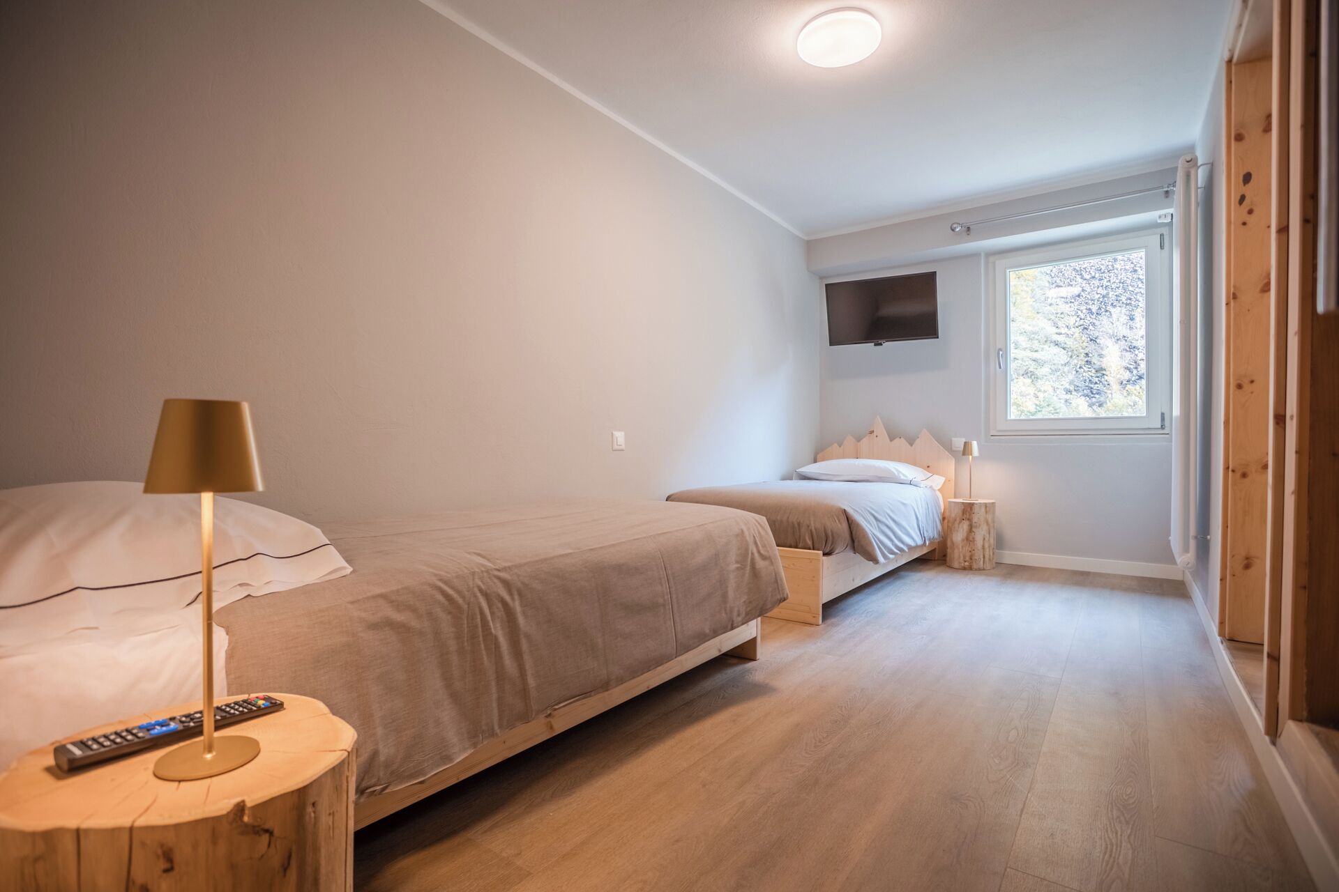Une chambre avec deux lits, un téléviseur fixé au mur et une fenêtre laissant entrer la lumière naturelle.