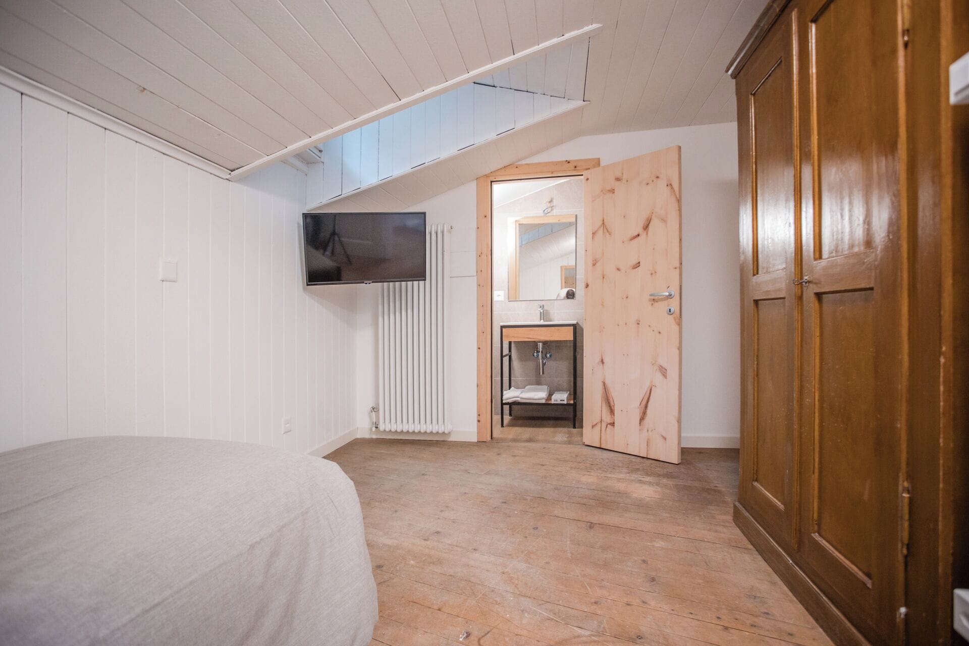 Een slaapkamer met een bed, een houten garderobe, een badkamer met een wastafel en spiegel, en een dakraam.