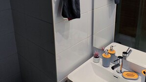 A modern bathroom with a white sink, mirror, and grey tiles. There are two towels hanging on towel rings, a toilet, and a paper towel dispenser. - Movistar Arena | National Stadium | Caupolicán (Santiago)