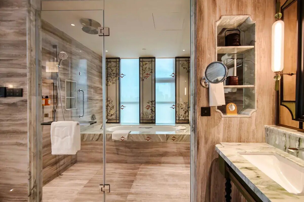 A modern bathroom with a glass shower enclosure, a bathtub, a vanity with a mirror, and a view of the city.