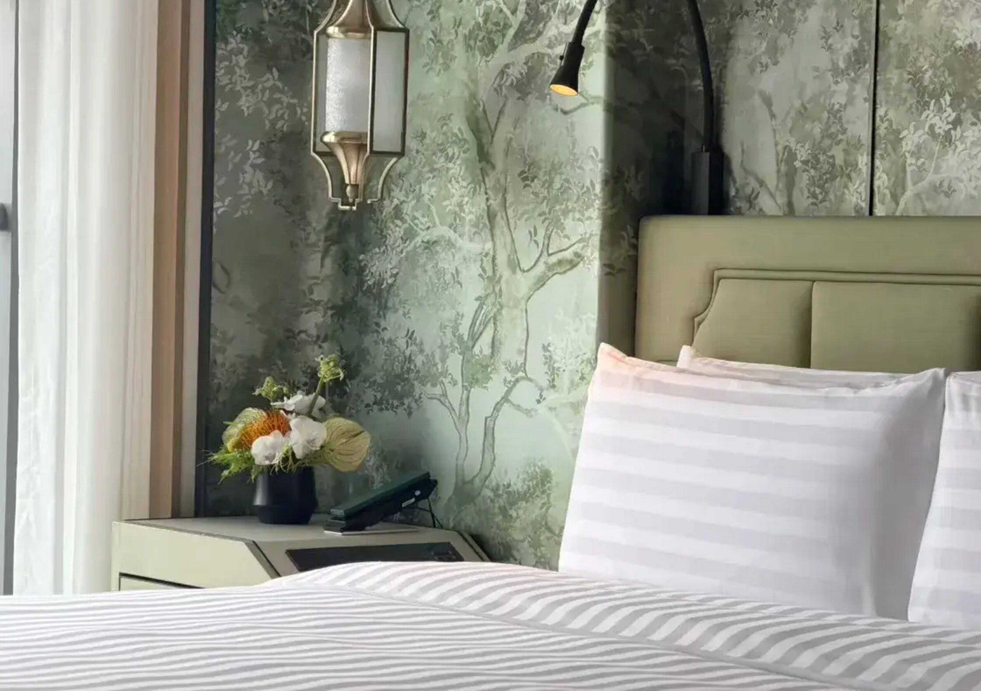 A bed with striped pillows, a nightstand with a floral vase, a lamp, and a wall with a tree pattern.