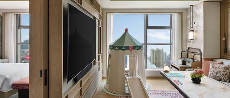 Habitación de hotel con una televisión de pantalla plana grande, una zona de juegos con un tobogán, una cama, un escritorio con lámpara y vistas a la ciudad.