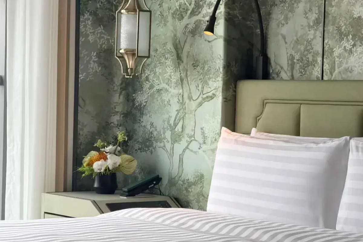 Un lit avec des coussins à rayures, une table de chevet ornée d’un vase à motifs floraux, une lampe et un mur décoré d’un motif représentant un arbre.