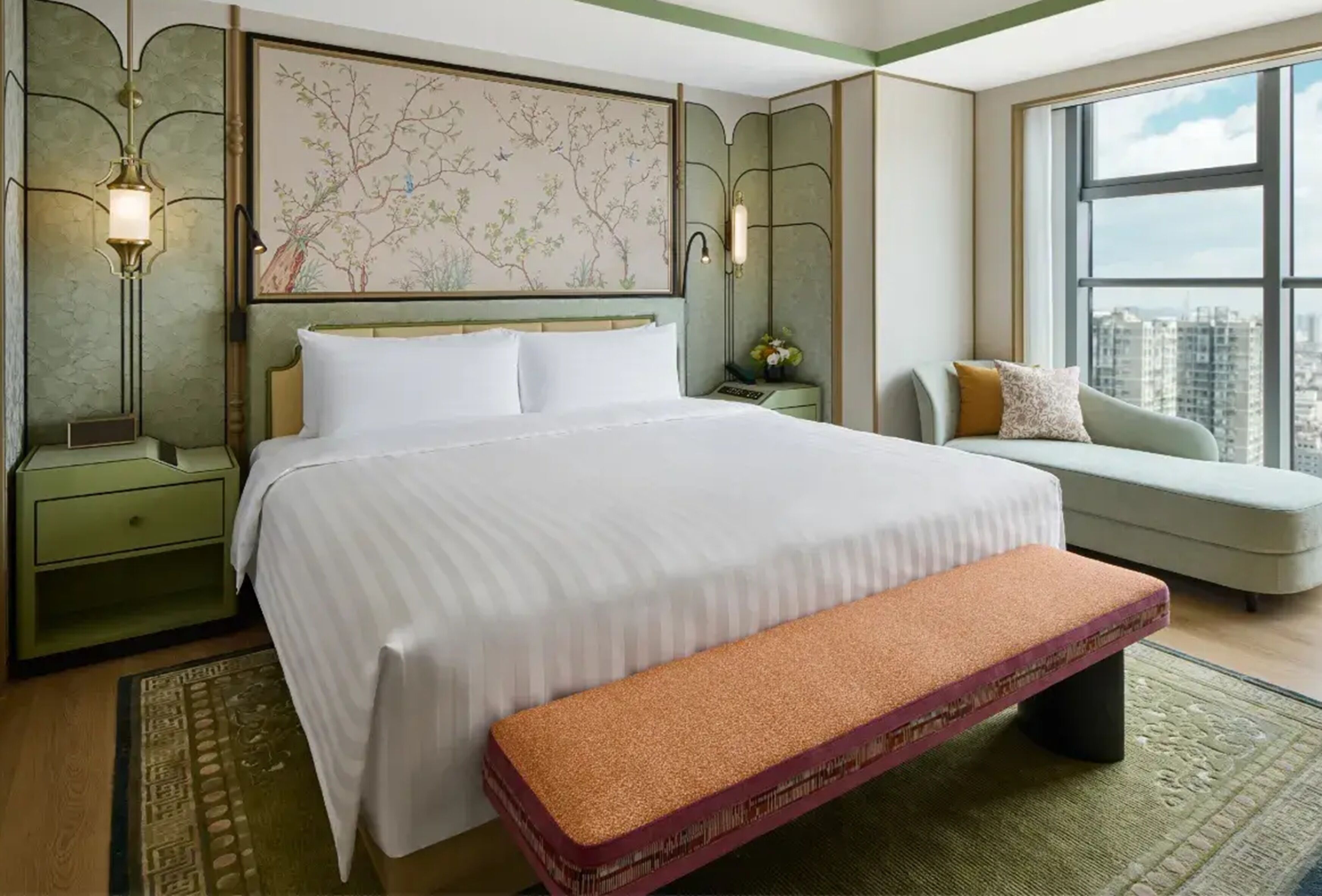 A hotel room with a large bed, a bench, a sofa, a nightstand, a wall-mounted lamp, and a window with a city view.