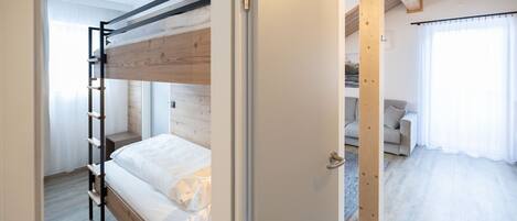 A bunk bed room with a wooden ladder, a door leading to a living area with a sofa and curtains.
