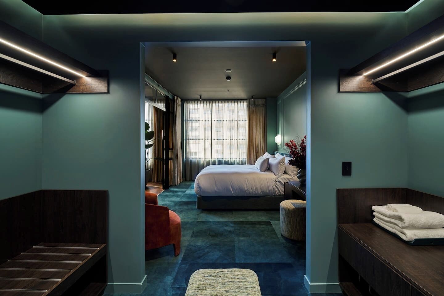 Habitación de hotel moderna con una cama grande, zona de estar con un banco y reposapiés, una mesita y vistas a la ciudad a través de amplios ventanales.