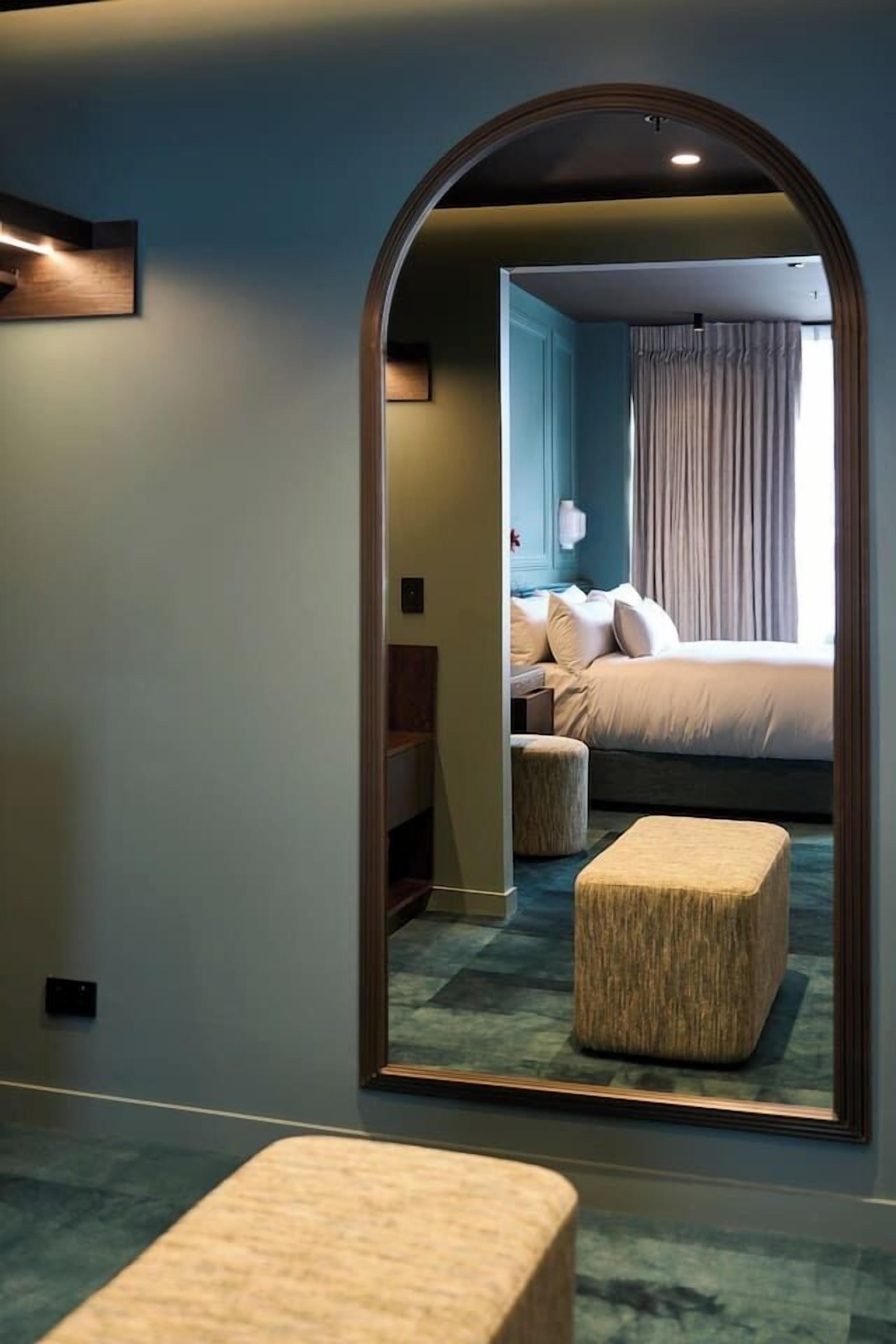 A hotel room with a large mirror reflecting the bed and curtains, a small ottoman, and a nightstand with a lamp.