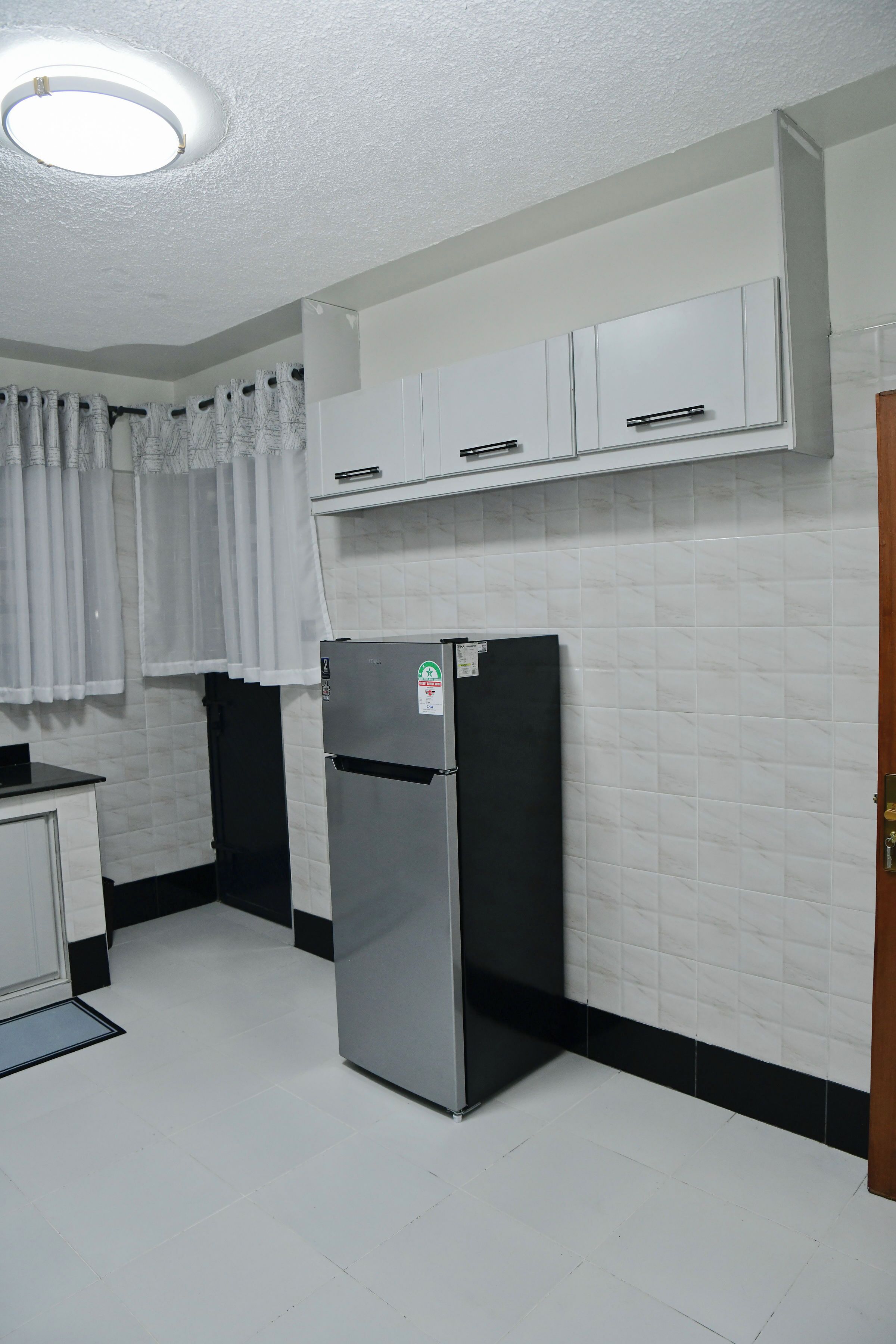 Una cocina con armarios blancos, un refrigerador negro y una ventana con cortinas blancas.