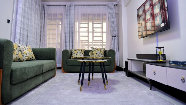 A living room with a green sofa, a small table, and a TV mounted on the wall. - Araka Homes-Naka/Exquisite 2 Bedroom Apartment (Nakuru)