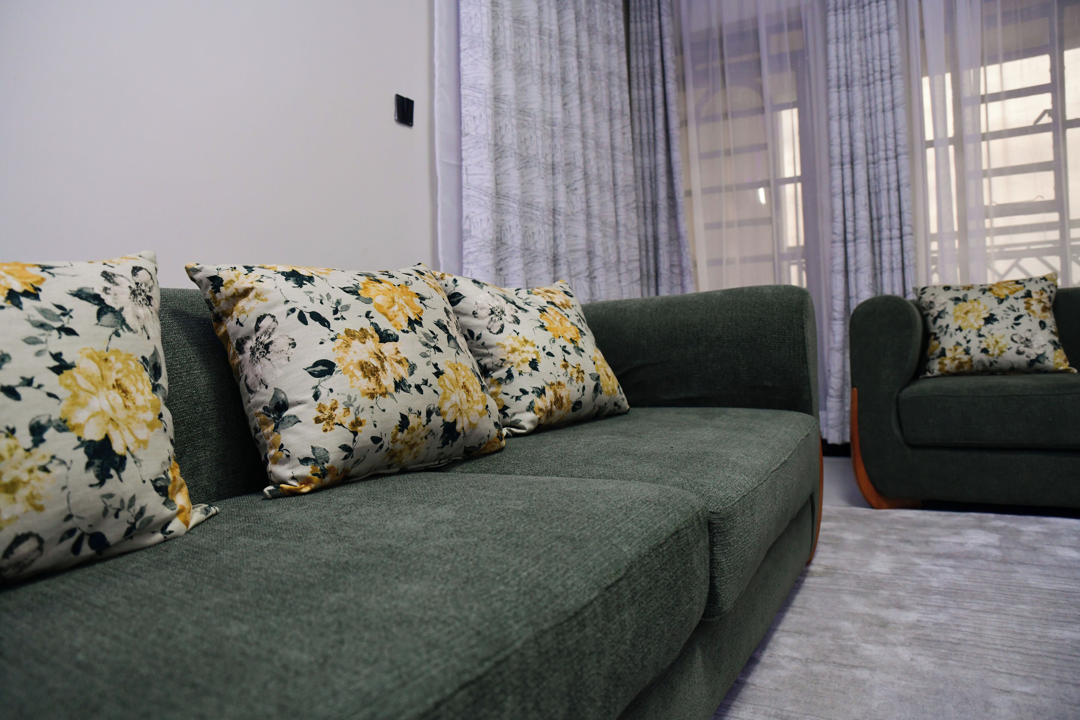 Un sofá verde con cojines estampados en una habitación con ventana y cortinas.
