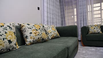 Un sofá verde con cojines estampados en una habitación con ventana y cortinas.