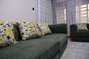 Een groene bank met bloemenkussens in een kamer met een raam en gordijnen.