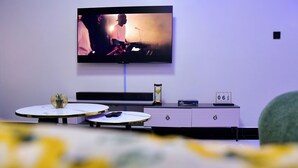 A modern living room with a flat-screen TV mounted on the wall, a small table with a decorative item, a clock, and a drawer unit. - Araka Homes-Naka/Exquisite 2 Bedroom Apartment (Nakuru)
