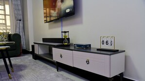A modern living room with a flat-screen TV mounted on the wall, a grey sofa, a small table, and a white console table with drawers. - Araka Homes-Naka/Exquisite 2 Bedroom Apartment (Nakuru)