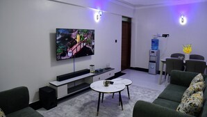 A modern living room with a flat-screen TV, a sofa, a coffee table, and a dining area with chairs and a water dispenser. - Araka Homes-Naka/Exquisite 2 Bedroom Apartment (Nakuru)