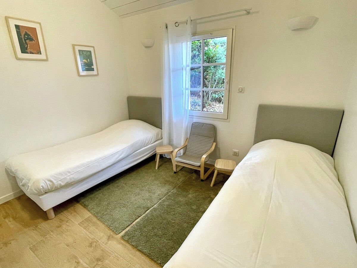 Una habitación con dos camas individuales, una silla pequeña, un ventanal con cortinas y una alfombra verde.