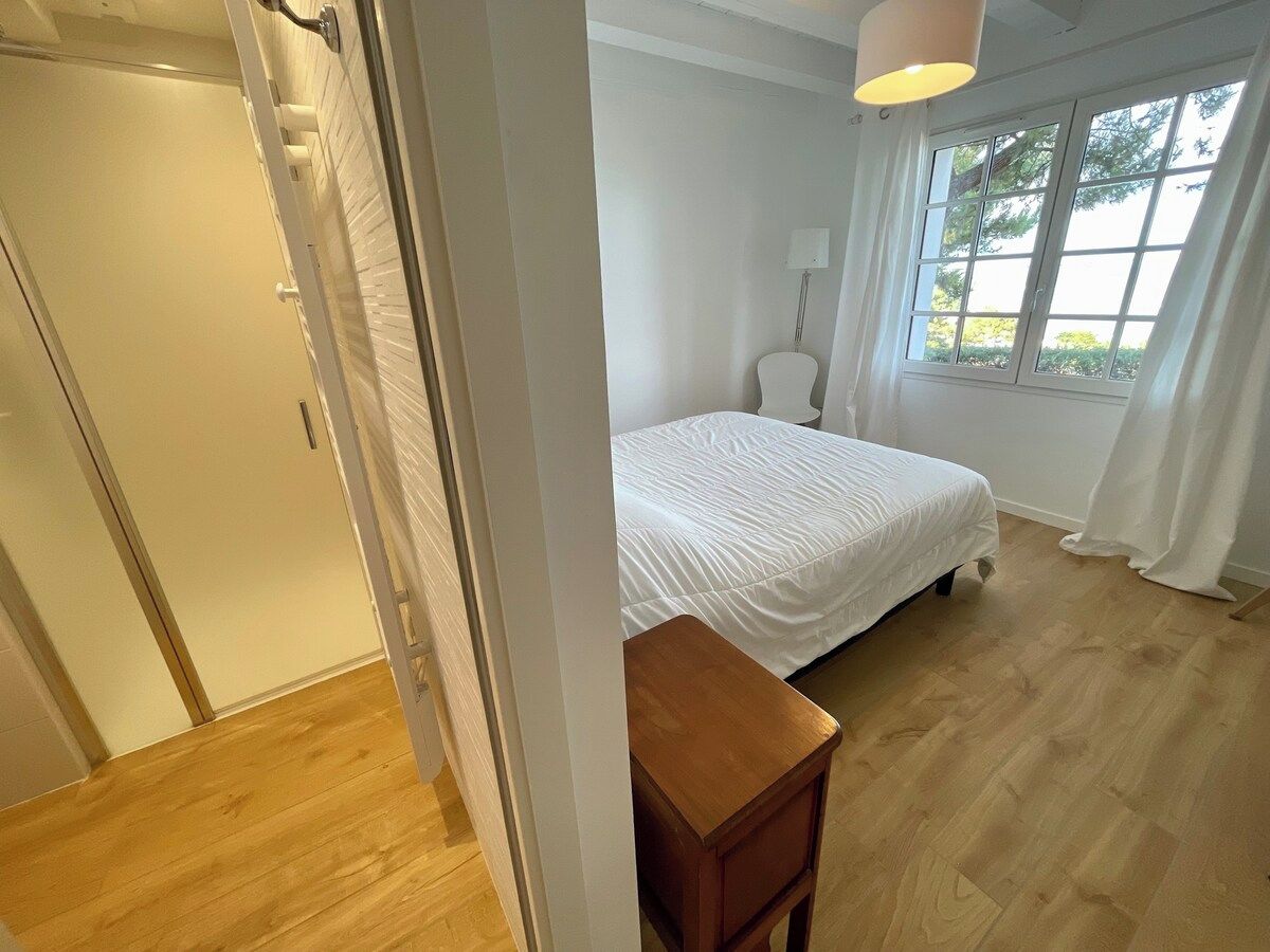 Un dormitori amb un llit, una taula de nit, una finestra amb cortines i una porta que dona a una altra habitació.